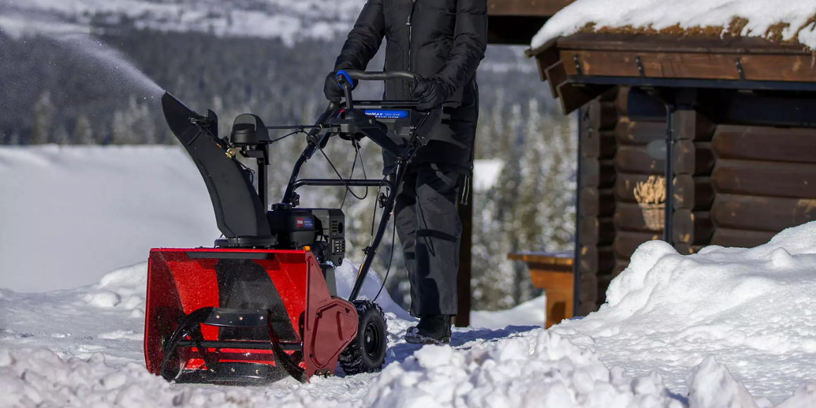 Dame i vinterklær og lue fjerner snø foran hytten med elektrisk snøfreser.