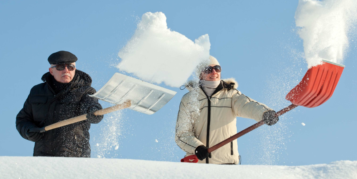 Eldre mann og ung kvinne i vintertøy måker tak med hver sin snøskuffe. I forkant ser vi snøen som kastes mot kamera.