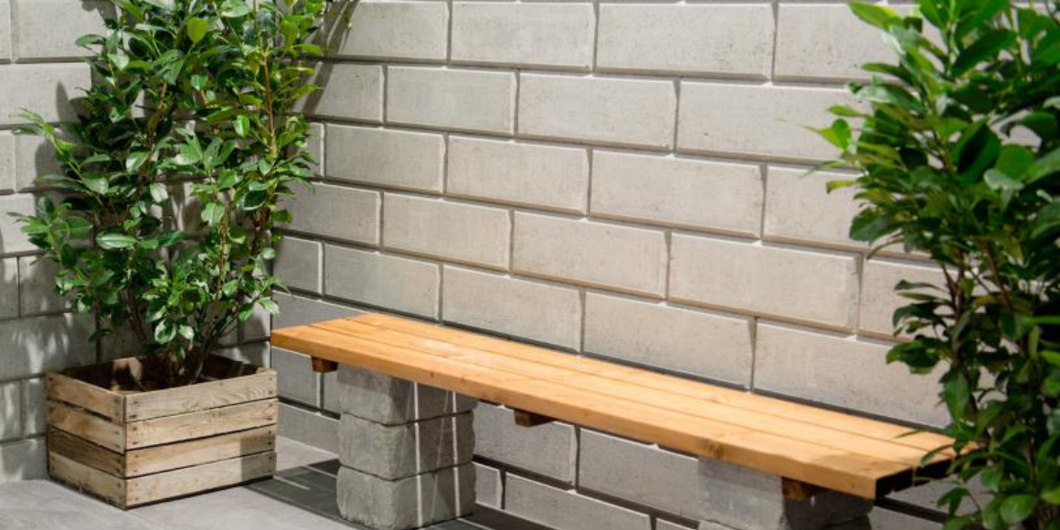 En minimalistisk utendørs setting med en trebenk støttet av Kombimur Støttemur, plassert mot en grå murvegg, flankert av to grønne potteplanter.