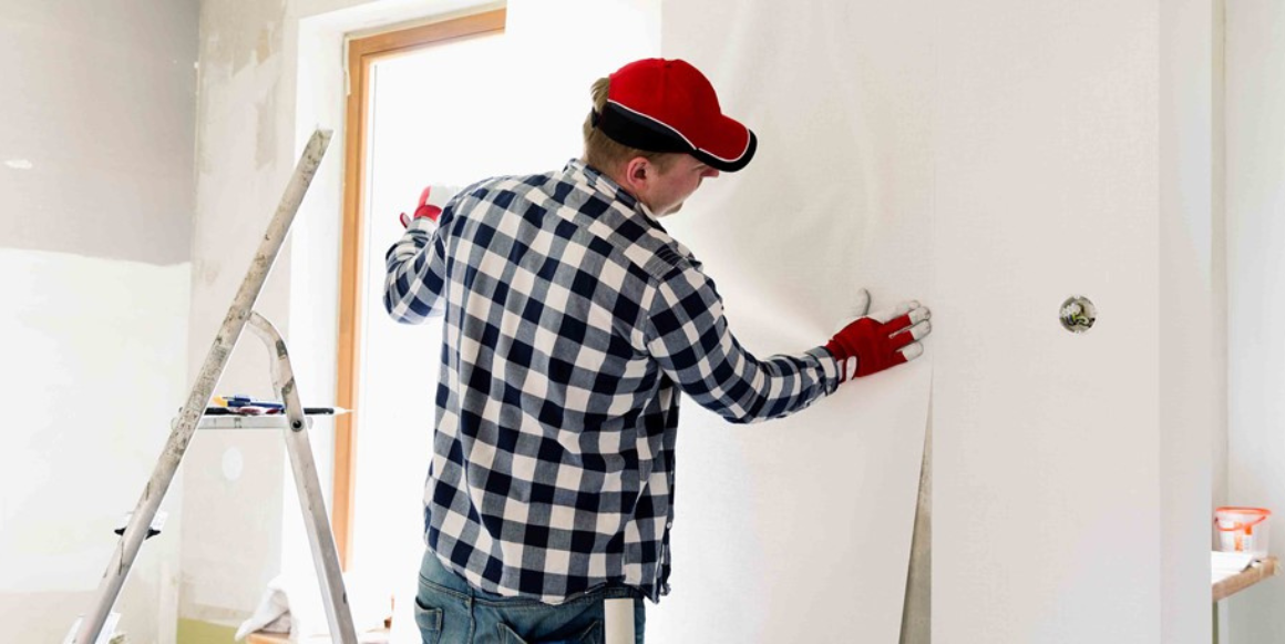 Mann med jeans, caps, ruteskjorte og hansker monterer gipsplater i et rom.