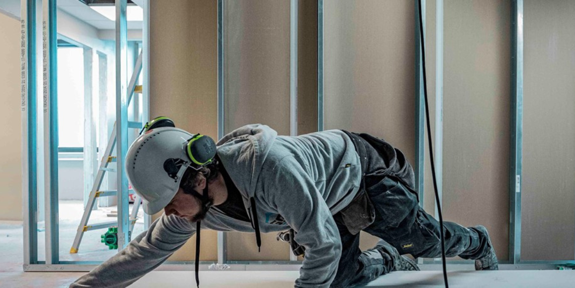 Håndverker på byggeplass lener seg over en stabel gipsplater for å måle og markere kappet på den øverste gipsplata.