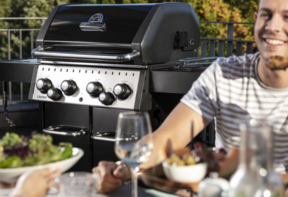 Mann som sitter ved et bord med sommermat og en stor gassgrill i bakgrunnen Mann som sitter ved et bord med sommermat og en stor gassgrill i bakgrunnen