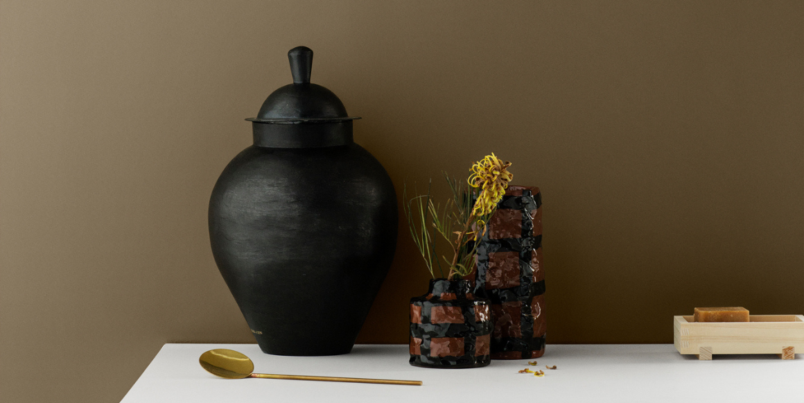 Dekor med mørk keramikkvase og naturmaterialer foran vegg malt i Jotun 1973 Golden Bronze.